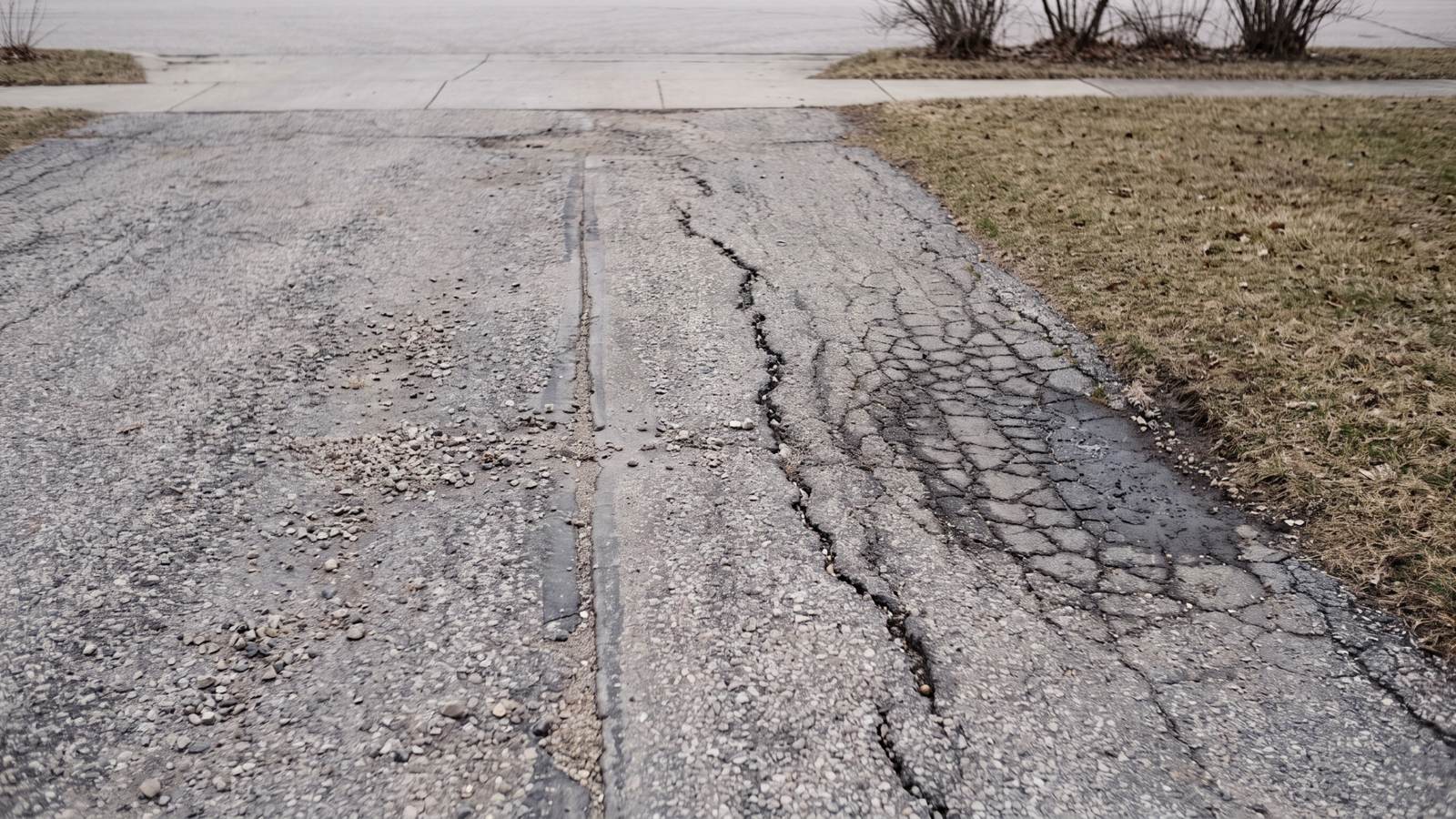 Dormant brown grass borders a worn driveway with raveled pebbles, a straight joint crack and a web of alligator cracking at its edge
